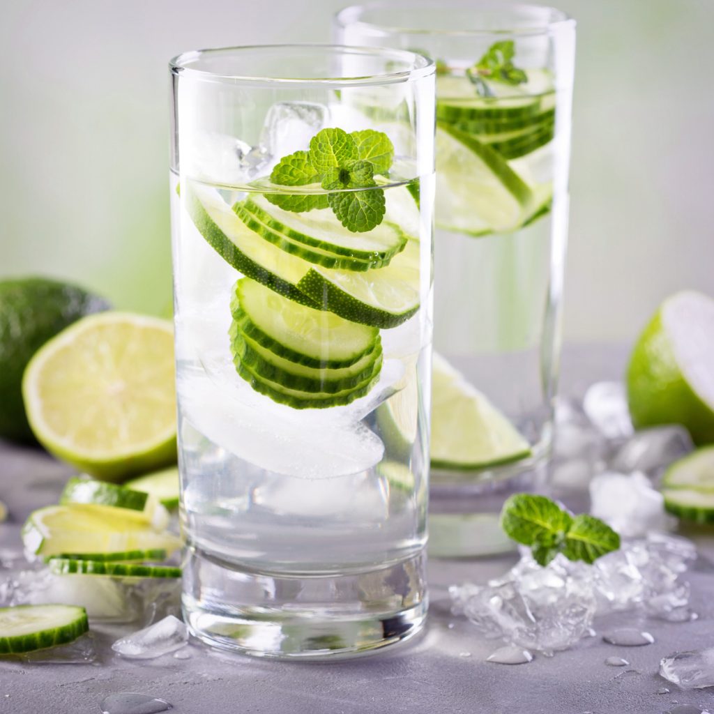 two glasses of cucumber slices in ice water with lime and cucumber slices around the glasses